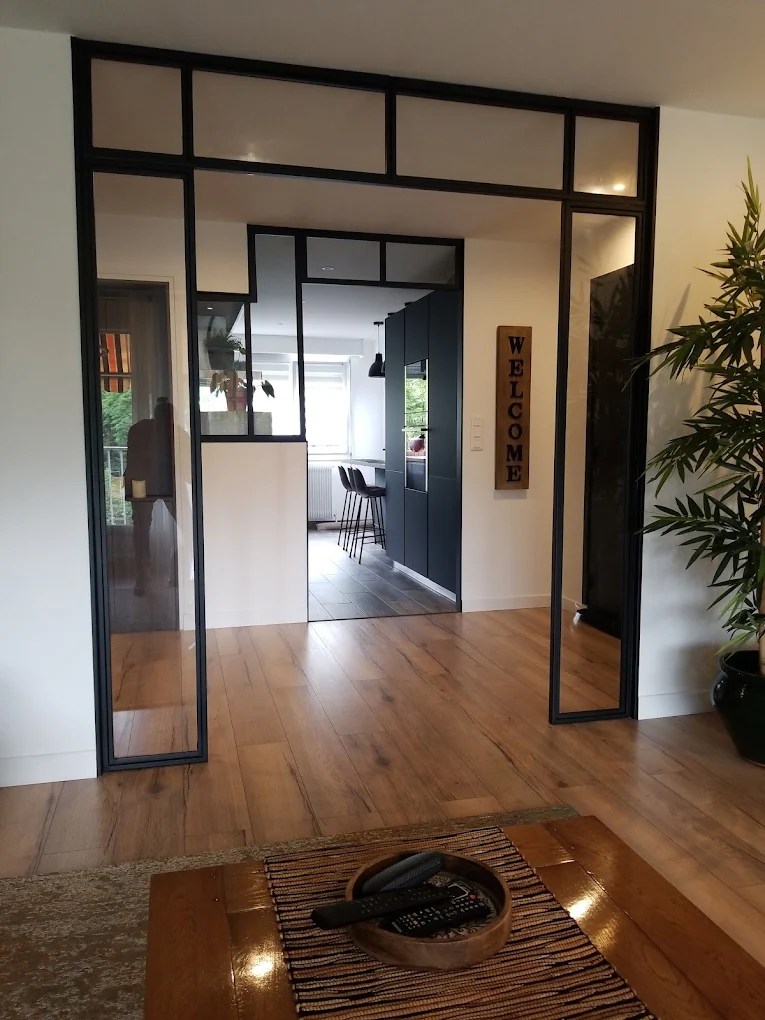 Vue d'un espace intérieur avec une verrière en métal séparant deux zones, comprenant un meuble de cuisine moderne et des chaises bar.